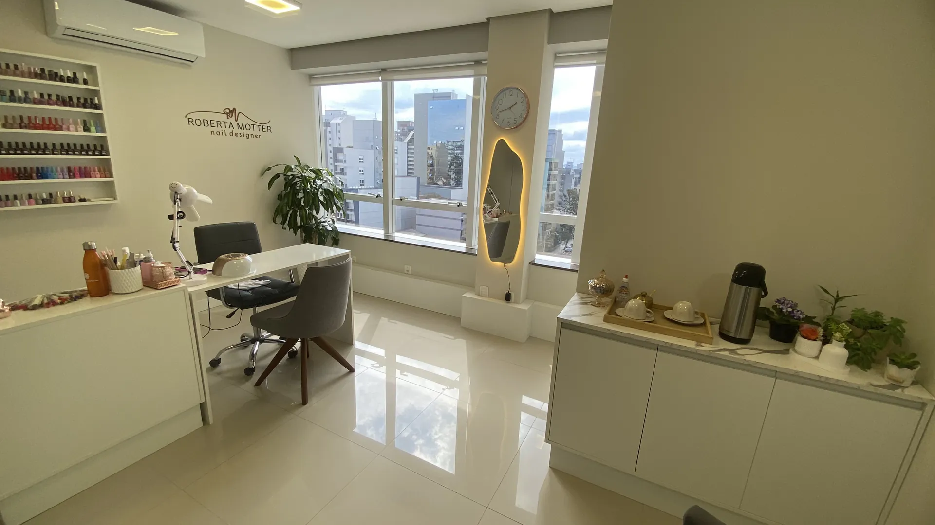 Sala do estúdio Roberta Motter: mesa de manicure clara, poltrona, plantas e prateleira de esmaltes junto a janelas amplas